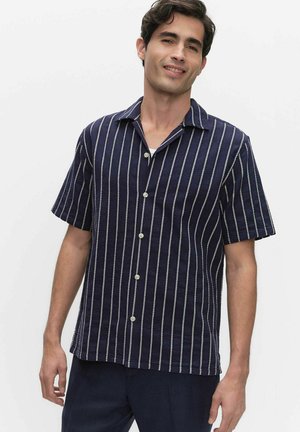 Homme portant une chemise à manches courtes bleu marine à boutons avec des rayures blanches verticales, debout devant un fond blanc uni.