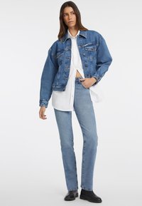 Giacca di jeans blu medio con grandi tasche frontali, chiusura a bottoni e una vestibilità corta, indossata sopra una camicia bianca a taglio dritto.