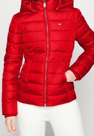 Red quilted puffer jacket with front zipper, side pockets, and small logo on chest, worn with white textured pants.