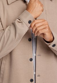 Giacca button-up beige con una texture morbida, bottoni neri e uno strato interno a righe visibile al colletto e ai polsini.