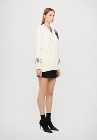 Bela pletena cardigan s kabelskimi vzorci in črnimi akcenti, s velikimi gumbi. Nosi jo čez črno mini krilo in črne pete.