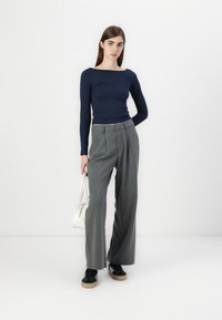 Top azul marinho de mangas compridas e calças cinzentas plissadas de cintura alta. Modelo segura uma mala branca, vestindo ténis pretos com sola castanha.