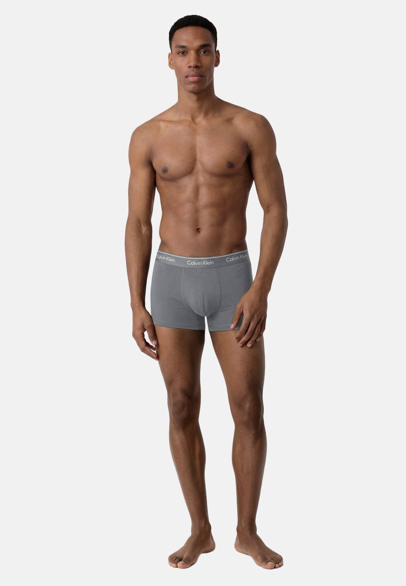 Homme musclé debout, pieds nus, portant un boxer gris Calvin Klein sur un fond clair uni.