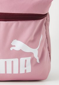 Mochila de tela rosa con un bolsillo lateral de malla, cremallera marrón y un gran logo blanco de Puma con el nombre de la marca impreso debajo.