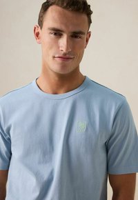 Lichtblauwe katoenen t-shirt met korte mouwen en een ronde hals. Bevat een klein geborduurd groen logo op de linkerkant van de borst. Zachte textuur.