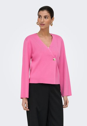 Femme portant un haut rose à manches longues avec un col en V et un bouton doré unique, associé à un pantalon noir taille haute, regardant sur le côté.