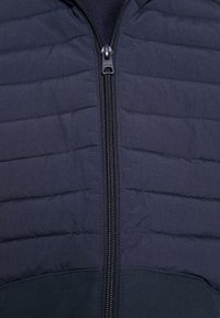 Marinblå puffjacka med dragkedja fram, med horisontell sömnad för textur och en slät tygfinish. Lättvikts- och minimalistisk design.