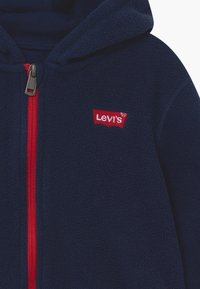 Granatowa bluza polarowa z pełnym czerwonym zamkiem, z czerwoną naszywką logo Levi's na klatce piersiowej oraz fakturowaną powierzchnią materiału.