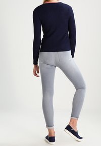 Jersey de punto azul marino, ajustado, con cuello redondo. Vaqueros pitillo gris claro con bajos remangados. Zapatos oscuros sin cordones con suela blanca.