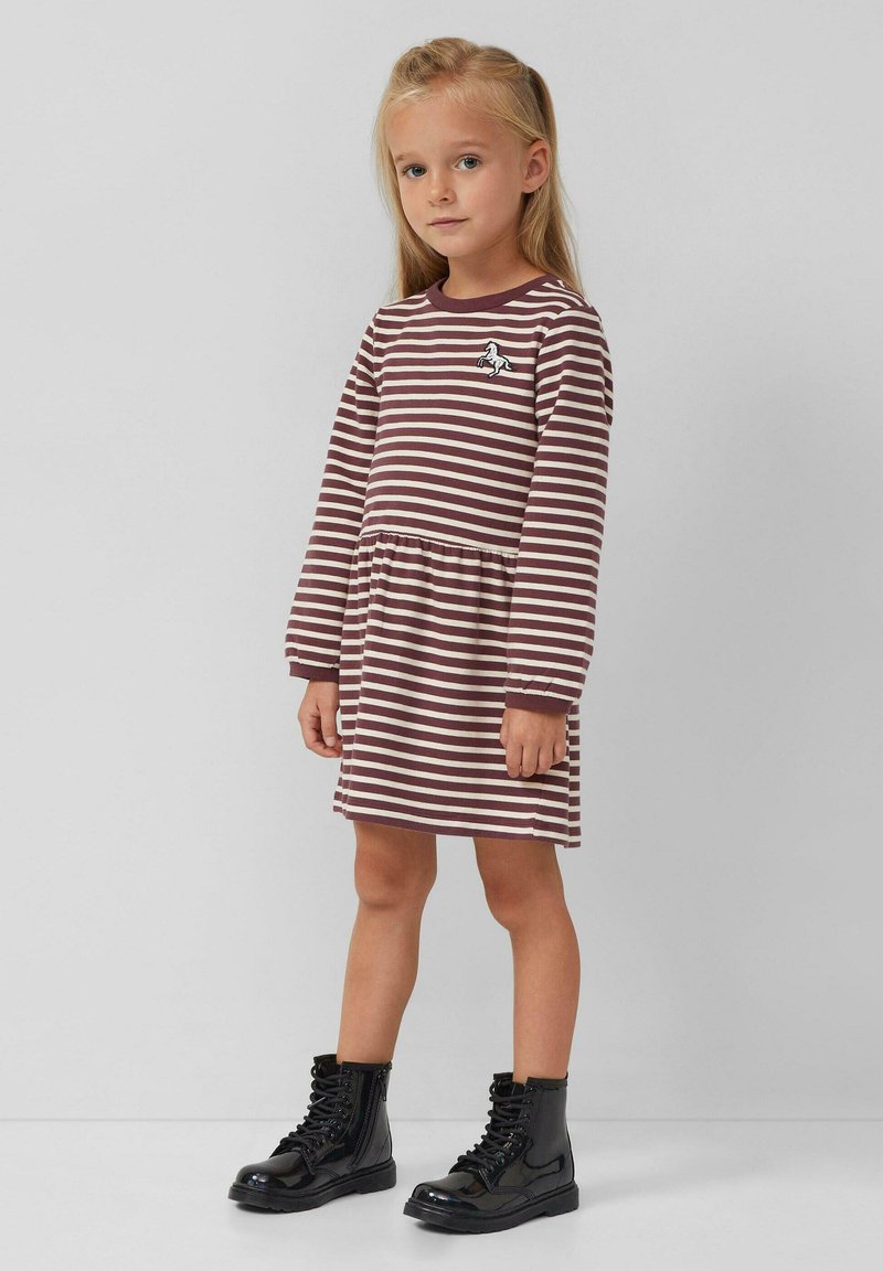 Sukienka w paski w kolorach brązowym i kremowym, z długimi rękawami, dopasowaną talią oraz zabawną naszywką jednorożca na piersi. Noszona z czarnymi butami typu combat.