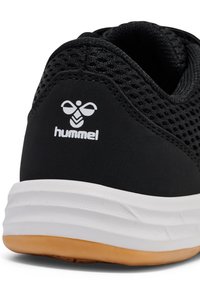 Talons d'une basket noire en tissu mesh, logo Hummel blanc, semelle intermédiaire blanche avec une semelle extérieure en caoutchouc marron clair.