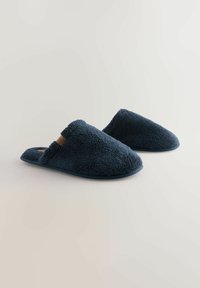 Paar von marineblauen, flauschigen Slippers zum Hineinschlüpfen mit niedrigen, offenen Rücken auf einer schlichten, hellen Oberfläche.