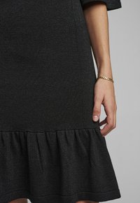 Svart klänning med texturerad tyg, med en utsvängd, volangkant. Ärmsluten är något bredare. Ett guldarmband syns på handen.