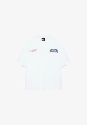 T-shirt blanc à manches courtes avec texte bleu marine "GOOD VIBES LAUNDROMAT" et texte rouge sur la poitrine, fond uni.