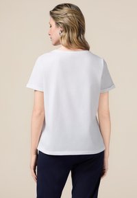 Camiseta de algodón blanca con mangas cortas, aberturas laterales en el dobladillo y un ajuste relajado. Textura suave con costuras mínimamente visibles.