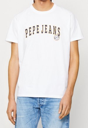 Osoba ubrana w białą krótką koszulkę z nadrukiem "PEPE JEANS" i niebieskie dżinsy stojąca na jednolitym jasnym tle.