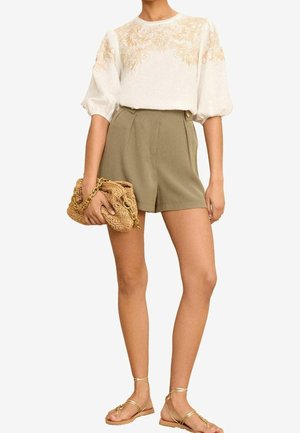Vrouw die een witte blouse draagt met beige bloemembroiders, olijfgroene shorts met hoge taille, een geweven clutch met gouden ketting vasthoudt en gouden sandalen met bandjes.