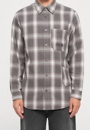 Homme portant une chemise à manches longues à carreaux gris et blanc avec deux poches avant et un pantalon gris.