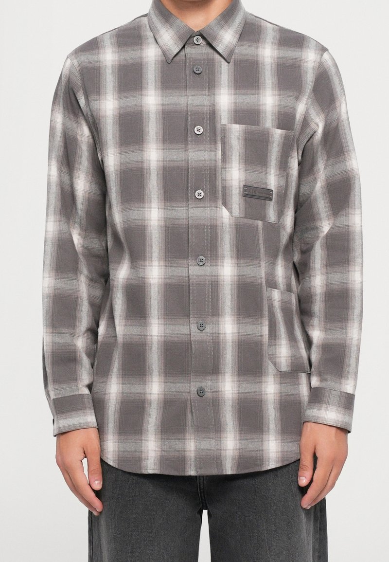 Homme portant une chemise à manches longues à carreaux gris et blanc avec deux poches avant et un pantalon gris.