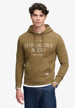 Mężczyzna ubrany w oliwkowo-zieloną bluzę z kapturem Superdry, niebieskie dżinsy i zegarek na nadgarstek, stojący na tle jednolitego białego tła.