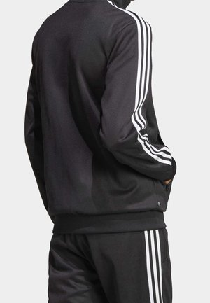 Homme portant un survêtement noir avec des bandes blanches sur les bras et les jambes, debout les mains dans les poches de la veste, vu de dos.