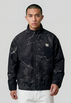 Junger Mann mit kurzem Haar trägt eine schwarze, marmorierte Jacke mit Reißverschluss, einem weißen Logo auf der Brust und weiße Hosen, steht vor einem schlichten Hintergrund.