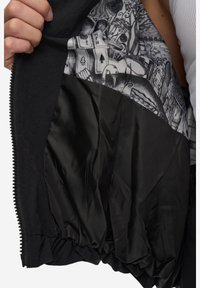 Černá bunda s texturovanou vnější vrstvou, s černou podšívkou připomínající hedvábí, zdobenou monochromatickými grafickými vzory. Otevírání na zip.