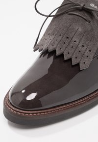 Chaussure en cuir verni noir avec détail frangé en daim gris, présentant des perforations, une forme ronde à l'avant, et une semelle contrastante marron cousue.