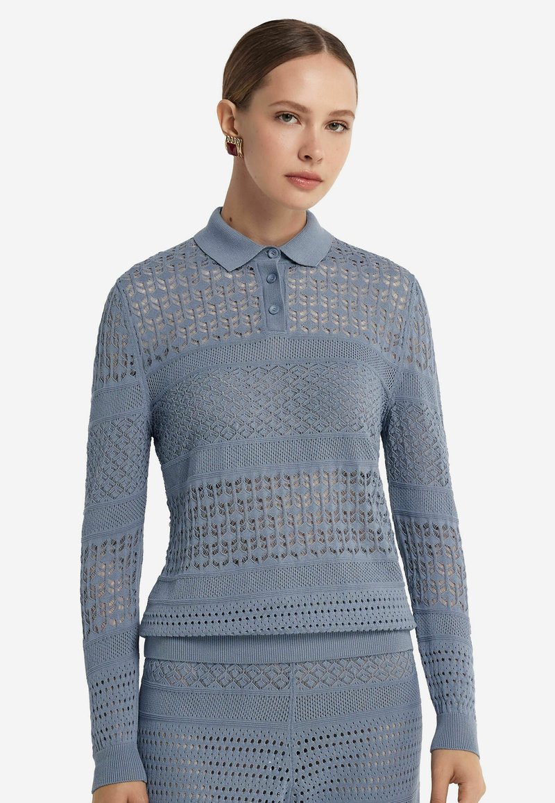 Ljusblå stickad långärmad polo med mönster, tre knappar, ribbad krage och manschetter. Texturerade sektioner skapar visuell kontrast.