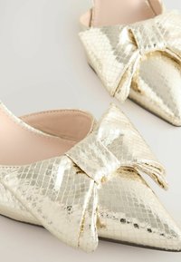 Ballerines dorées avec un design à bout pointu, ornées d'un grand nœud et d'une finition métallique texturée. Doublure intérieure douce et semelle plate.