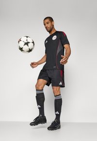 Sort atletisk trøje med røde accenter, sorte shorts, sorte sokker med hvide striber og sorte fodboldstøvler; en fodbold svæver i nærheden.