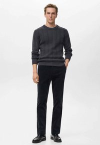 Suéter de punto acanalado en gris oscuro con cuello redondo, combinado con pantalones de pana negros de corte ajustado y zapatos negros, de pie contra un fondo claro.