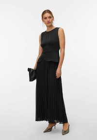 Femme portant un haut noir sans manches et une jupe longue noire plissée, tenant une pochette noire, chaussant des talons aiguilles scintillants et des boucles d'oreilles pendantes.