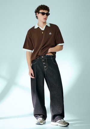 Junger Mann mit dunkler Sonnenbrille, braunem kurzärmligem Poloshirt, weitem dunklem Jeans, Turnschuhen und einer Anhängerkette, stehend vor einem hellen Hintergrund.