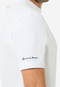 Weißes Poloshirt mit geripptem Kragen, kurzen Ärmeln und einem navyblauen gestickten Logo auf dem linken Ärmel. Strukturierte Stoffoberfläche.