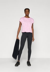 Απαλό ροζ κοντομάνικο τοπ με λεπτό μοτίβο λογότυπου, συνδυασμένο με μαύρα κολάν και μαύρα αθλητικά παπούτσια, κρατώντας μια μαύρη πετσέτα.