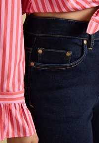 Gros plan sur un jean en denim bleu foncé avec des coutures visibles et des rivets métalliques à côté d’une personne portant une chemise rayée rose et rouge.