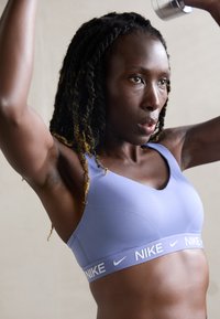 Mujer con trenzas que lleva un sujetador deportivo morado de Nike levantando mancuernas por encima de su cabeza, concentrada durante una sesión de entrenamiento de fuerza.