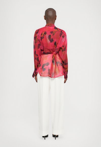 Translucente roze blouse met bloempatroon, lange mouwen en een strik in de taille, gecombineerd met witte wijde broekspijpen.