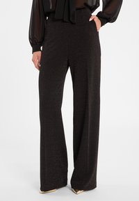 Pantalon large noir avec une texture scintillante, dotés d'une taille haute et de poches latérales, associé à un chemisier noir transparent.