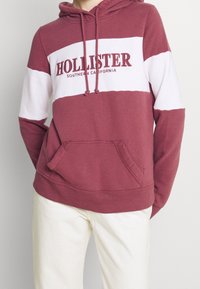 Persona con una sudadera color burdeos con rayas blancas y el texto "HOLLISTER Southern California", combinada con pantalones color crema.