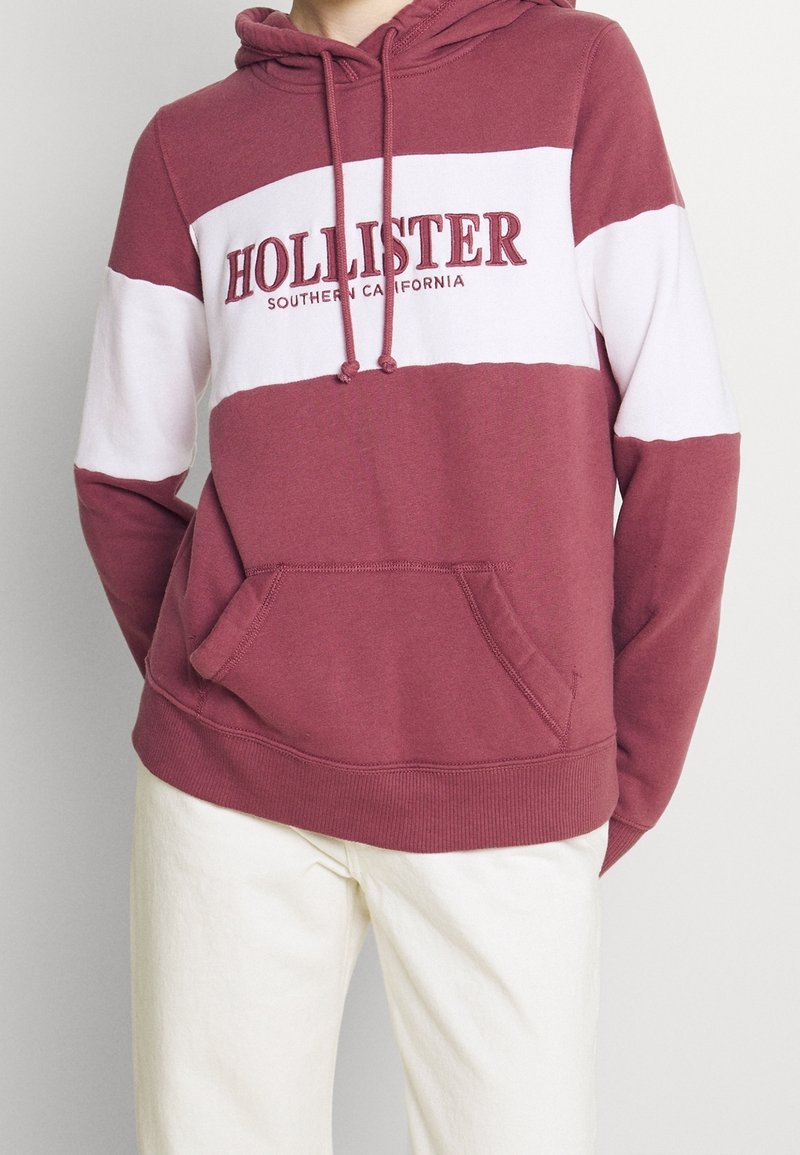 Persona con una sudadera color burdeos con rayas blancas y el texto "HOLLISTER Southern California", combinada con pantalones color crema.