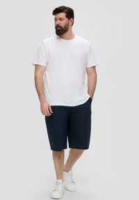 T-shirt en coton blanc, shorts navy et baskets blanches. Le modèle se tient dans une pose détendue, mettant en avant une tenue décontractée au design simple.