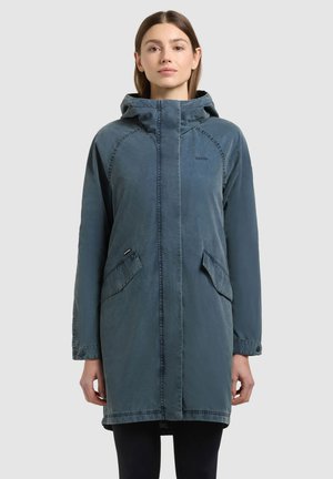 Nő, aki hosszú, kék kapucnis dzsekit visel elülső zsebfedőkkel, egyszerű világosszürke háttér előtt áll.