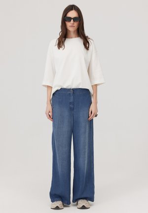 Kobieta w czarnych okularach przeciwsłonecznych, luźnej białej bluzce o długości rękawa za łokieć, szerokich niebieskich jeansach i beżowych sneakersach, stojąca na jednolitym tle.