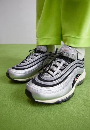 Osoba nosząca srebrno-czarne buty sportowe Nike z różowymi akcentami, białe skarpetki oraz jaskrawozielone spodnie stojąca na zielonym dywanie.