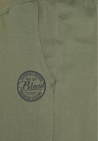 Tissu vert olive avec une texture lisse, présentant une poche sur le côté gauche et un logo circulaire bleu marine avec le texte "Blend" et des détails de qualité premium.