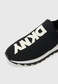 Μαύρο αθλητικό παπούτσι χωρίς κορδόνια με υφή στο πάνω μέρος, έντονο λευκό λογότυπο, στρογγυλεμένη μύτη και σόλα με πολυκατευθυντικό σχέδιο.