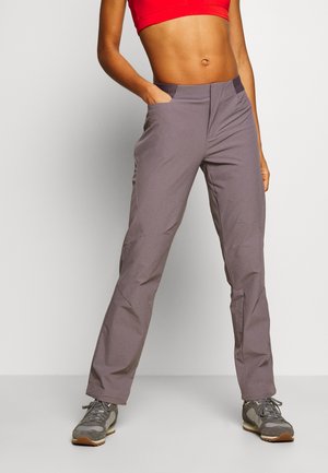 Mujer con pantalones grises y zapatillas grises, de pie con una mano en el bolsillo y top corto rojo sobre fondo liso.