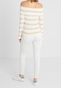 White ribbed off-the-shoulder top with orange stripes, paired with white skinny jeans and white sneakers, featuring green accents on shoes.